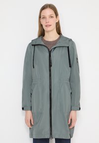 Femme portant un imperméable gris à capuche mi-long avec une fermeture éclair noire, des poches à l'avant et des poignets réglables, debout devant un fond blanc.