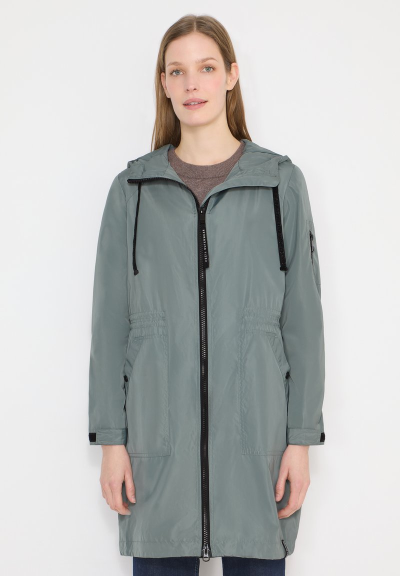 Femme portant un imperméable gris à capuche mi-long avec une fermeture éclair noire, des poches à l'avant et des poignets réglables, debout devant un fond blanc.