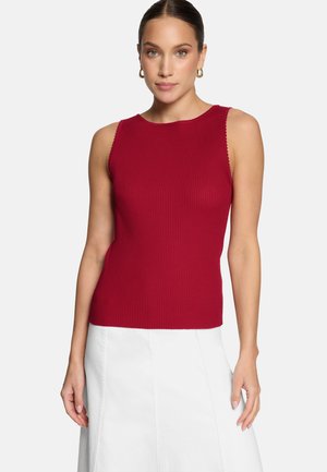 Femme portant un haut rouge côtelé sans manches et une jupe blanche, cheveux attachés, debout devant un fond blanc uni.