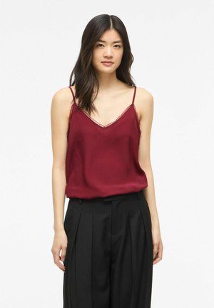 Femme portant un haut bordeaux sans manches à col en V avec bordure en dentelle et un pantalon plissé noir taille haute sur un fond uni.
