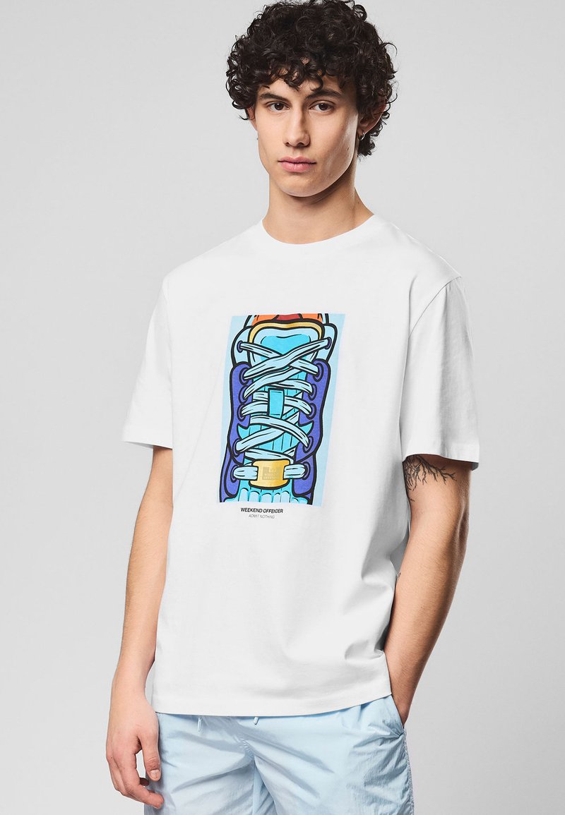 Jeune homme aux cheveux bouclés portant un t-shirt blanc avec un motif de basket colorée et un short bleu pâle, debout devant un fond uni.