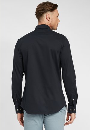 Man die met zijn rug naar de camera staat, gekleed in een strakke zwarte blouse met lange mouwen en knopen, en lichtblauwe broek, tegen een effen achtergrond.