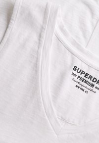 Débardeur blanc en coton avec col en V et texte de marque imprimé en noir "Superdry Premium" et "New York, N.Y." sur la partie intérieure du col.