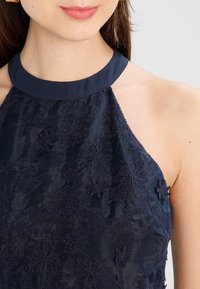 Navy sleeveless top with a halter neck design, featuring textured floral embroidery and a smooth satin finish.
