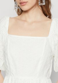 Biele šaty so štvorcovým výstrihom, vzorom broderie anglaise, balónovými rukávmi a zberaným pásom. Veľké, štvorcové kryštálové náušnice.