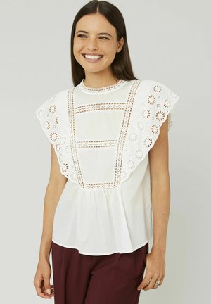 Blouse en coton blanc avec un bord en dentelle brodée et des manches bouffantes, dotée d'un col rond et d'un bas plissé. Associée à un pantalon bordeaux.