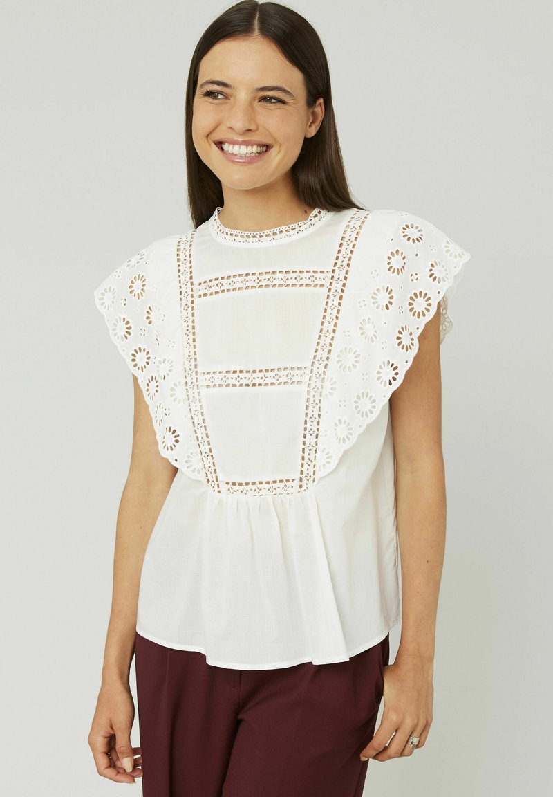 Blouse en coton blanc avec un bord en dentelle brodée et des manches bouffantes, dotée d'un col rond et d'un bas plissé. Associée à un pantalon bordeaux.
