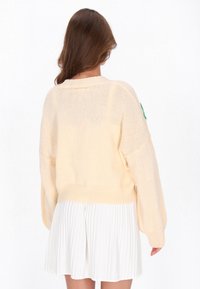 Jasno brzoskwiniowy sweter z dzianiny o luźnym kroju, z prążkowanym wykończeniem i zielonym detalem naszytym na rękawie, zestawiony z plisowaną białą spódnicą.