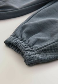 Grå atletikbukser har et tekstureret stof med elastiske manchetter, der er samlet, og viser en glat overflade og et åndbart design.