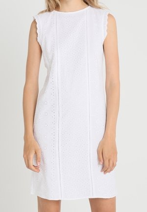 Femme portant une robe blanche sans manches en tissu ajouré avec broderies florales et à pois, posant devant un fond uni.