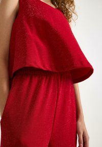 Rød ærmeløs buksedragt med en løs, draperet top og elastisk talje. Materialet har en subtil glans og en glat tekstur.