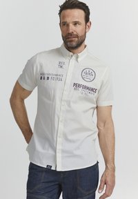 Camisa blanca de manga corta con cuello abotonado, que presenta texto y logos bordados en azul marino y púrpura, y una pequeña etiqueta en el dobladillo.