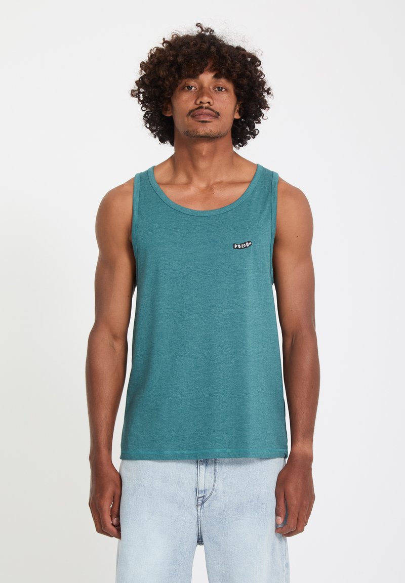 Homme aux cheveux bouclés portant un débardeur bleu sarcelle et un jean bleu clair, debout devant un fond blanc uni.