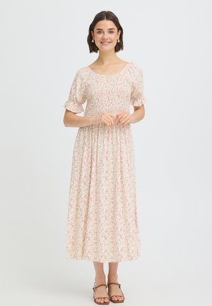 Młoda kobieta stoi, ubrana w jasną kwiatową sukienkę midi z bufkami i brązowe sandały na pasku przy kostce, uśmiecha się na jednolitym tle.