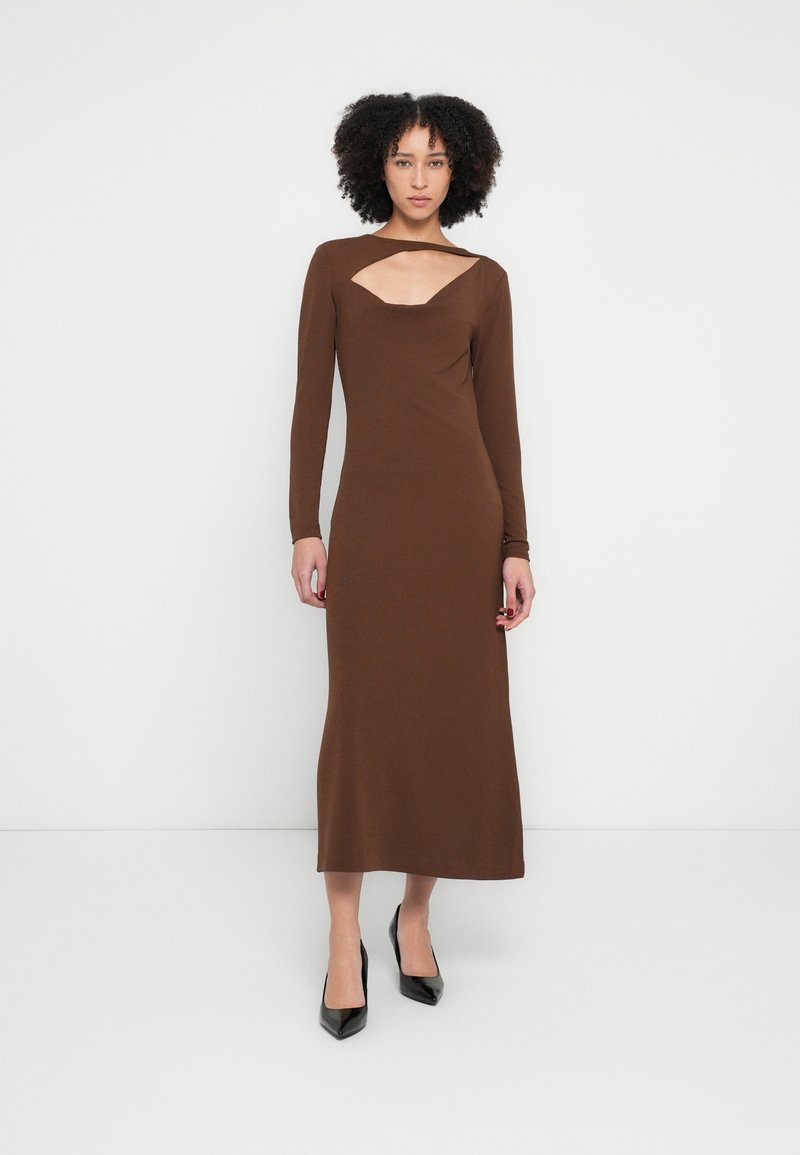 Brūna kleita ar garām piedurknēm un izliekta izgriezuma detaļu krūšu daļā. Izgatavota no gludas auduma, krīt līdz potītēm un kombinēta ar melniem piespraustiem papēžiem.