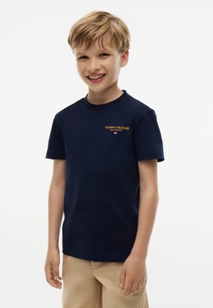 Smilende ung dreng med lyst brunt hår iført en marineblå Tommy Hilfiger T-shirt og beige bukser mod en ensfarvet hvid baggrund.
