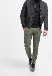 Černá bomber bunda přes černý rolák, v kombinaci s olivově zelenými kalhotami a černými teniskami. Texturovaná tkanina a přiléhavý střih.