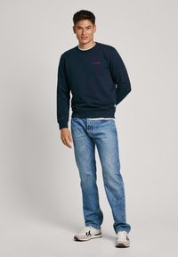 Felpa blu navy con logo rosso, colletto rotondo, maniche lunghe e polsini a coste. Abbinata a jeans azzurri e sneaker bianche.