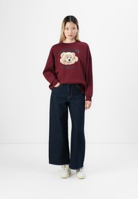 Young woman standing with hand in pocket, wearing a burgundy sweatshirt with teddy bear graphic and navy wide-leg jeans with white sneakers.