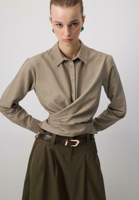 Beige-paitainen paita, jossa on kaulus ja kietaisusuunnittelu sekä teksturoitu kangas. Ruskea vyö kullanvärisellä soljella yhdistettynä tummanvihreisiin laskostettuihin housuihin.