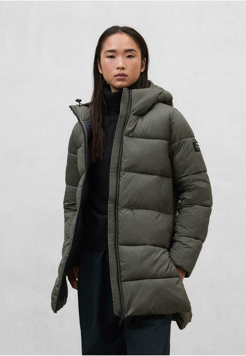 Mujer vestida con un abrigo largo acolchado de color verde oliva con capucha, un suéter negro de cuello alto y pantalones oscuros, de pie frente a un fondo claro y liso.