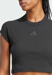 Black ribbed crop top with short sleeves, featuring a textured finish and tonal Adidas logo on the chest. Round neckline with subtle seams.