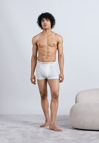 Young man with curly hair standing barefoot on gray carpet, wearing white Calvin Klein boxer briefs next to a rounded beige armchair.