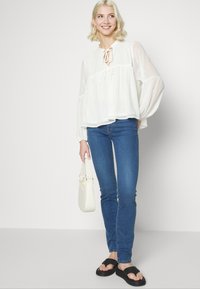 Blusa branca leve com mangas em balão e decote com laço, combinada com jeans skinny azuis e sandálias pretas de slide. Pequenos pontos decorativos na blusa.