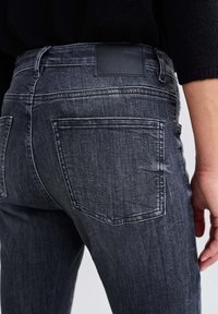 Jeans in denim grigio scuro con finitura testurizzata, due tasche posteriori e una patch in pelle nera sulla vita. Design minimalista.