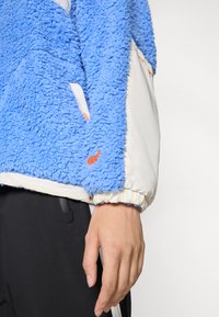 Fleecejakke med blå sherpa ytterstoff og hvite nylonærmer, med elastiske mansjetter og en oransje logo på ermet.