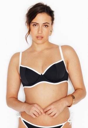 Donna con capelli scuri raccolti, che indossa un bikini nero con bordi bianchi, orecchini a cerchio dorati e braccialetti d'oro al polso.