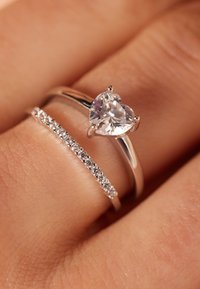 Twee zilveren ringen op een vinger: de ene heeft een hartvormige heldere edelsteen, terwijl de andere een rij kleine heldere stenen in een band heeft gezet.