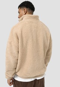 Beige fleece genser med høy krage, teksturert overflate, og avslappet passform. Har ribbestrikkede mansjetter og midje. Ingen synlig maskinvare.