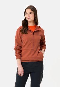 Pullover in pile di colore ruggine, con zip e collo alto, tessuto strutturato e tasche laterali, indossato sopra una maglietta arancione.