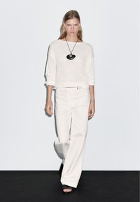 Witte gebreide trui met een cropped ontwerp, gecombineerd met hoge, wijde witte broeken. Heeft een grote zwarte hangerketting.