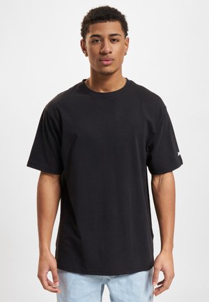 Jeune homme aux cheveux courts et bouclés, portant un t-shirt noir ample et un jean bleu clair, debout devant un fond blanc uni.