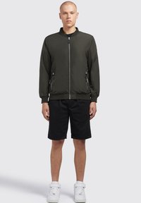 Sötétzöld bomber dzseki, cipzárral elöl, bordás gallérral és mandzsettákkal. Fekete rövidnadrággal és fehér magasszárú sneakers-szel párosítva.