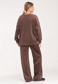 Brązowy dres z weluru, składający się z bluzki z długim rękawem i luźnych spodni, charakteryzujący się miękką teksturą i swobodnym krojem. Brak wyróżniających się detali.