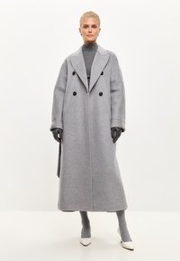 Palton lung din lâna gri, cu design dublu-botonat, nasturi black, rever mari și slituri laterale, purtat peste un pulover cu guler înalt și mănuși.