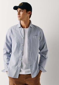 Camicia a righe blu e bianche con taschino, indossata sopra una maglietta bianca, abbinata a pantaloni marroni e un cappellino blu navy.