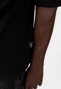 Černé tričko s krátkým rukávem z bavlněné tkaniny, se štítkem s logem na straně. Design je jednoduchý, s volným střihem a rovnými okraji.