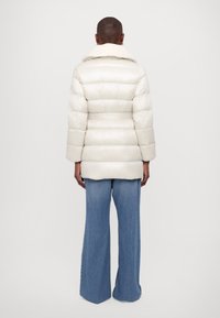 Puffer jacket in shiny cream material with a fur-lined collar, featuring a belted waist and quilted texture. Paired with wide-leg blue jeans.
