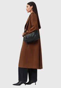 Borsa nera in pelle con dettagli zip dorati, caratterizzata da una texture morbida e una forma strutturata, indossata sotto un cappotto marrone.