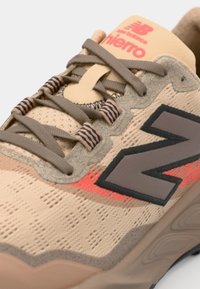 Baskets de sport beiges avec une tige texturée, des accents marron foncé, des détails rose-orange et des lacets plats. Présente un logo proéminent et un embout renforcé.