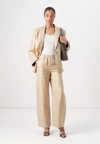 Blazer beige ajusté sur un débardeur blanc, associé à un pantalon large taille haute beige et des talons strappy noirs. Tissu léger, texture douce.