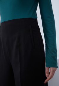 Lähivõte inimesest, kes kannab musti kõrge vöökohaga pükse külje luku ja türkiissinise pika varrukaga soonikkoega pluusiga, käsi lõdvestunult külje kõrval.