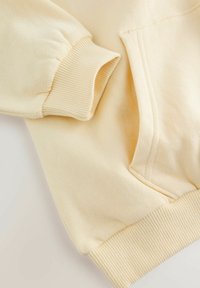 Krēmkrāsas džemperis ar aprocēm un apakšmalu ar ribotu audumu, uz vienkārša gaiša fona.