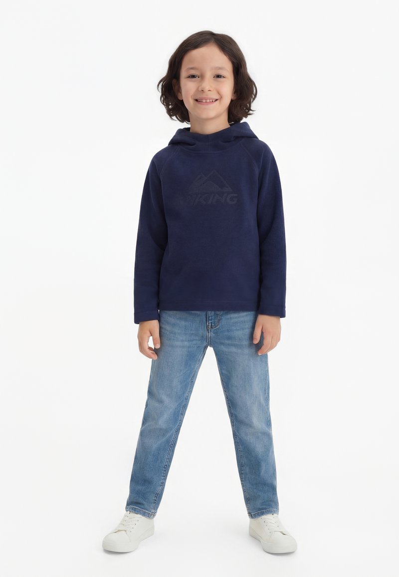 Naeratav laps, kellel on õlgadeni juuksed, kellel on seljas tume sinine kapuutsiga dressipluus, helesinised teksad ja valged tossud, seisab valgel taustal.