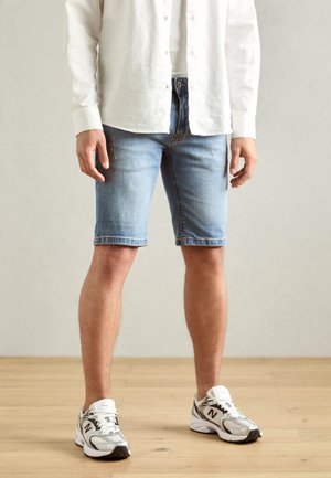 Shorts en denim de couleur bleu clair, longueur mi-cuisse avec un ourlet effiloché. Associés à une chemise blanche à boutons et des baskets gris-blanc.