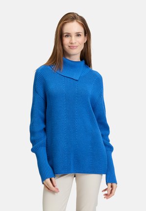 Blue knitted sweater featuring a high collar, textured fabric, wide sleeves, and a relaxed fit. Paired with light-colored pants.